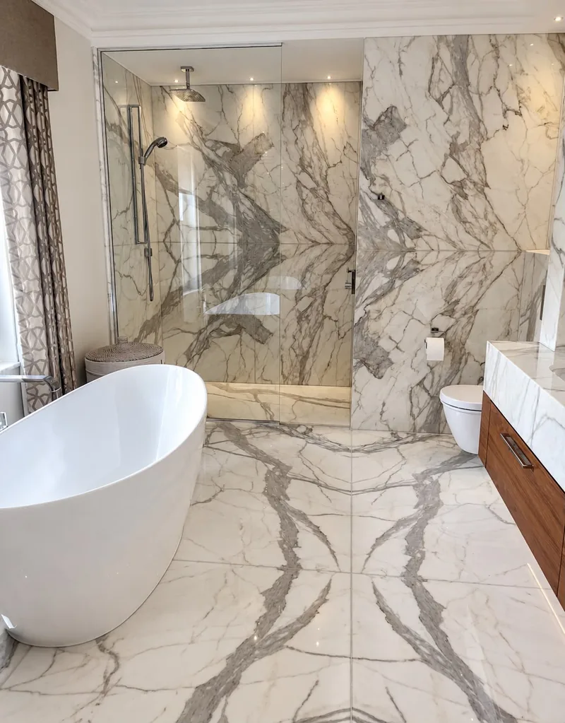 A bathroom lined with bookmatched white marble with grey veining on the floor and walls, a walk-in glass shower and a freestanding bath